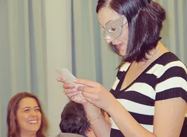 A woman in a masquerade mask reading a card while a guest laughs