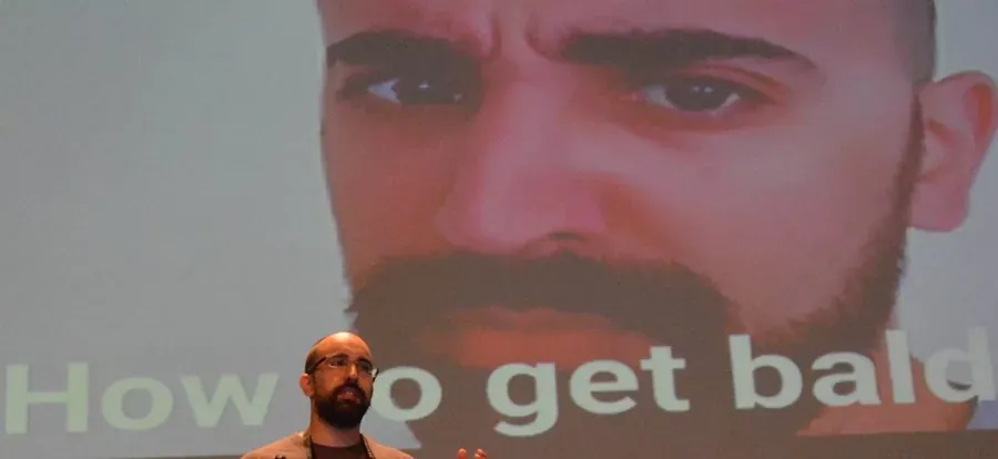 A speaker on stage beneath a giant projected close-up of a bearded face and the words “How to get bald”