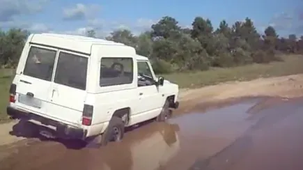 A white off-road jeep stuck in a muddy puddle on a dirt track, with trees behind it