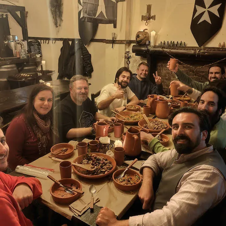 A group of friends at a long wooden table in a medieval-themed tavern, with clay jugs and bowls