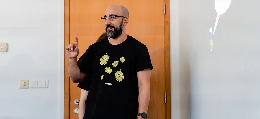 A speaker in a black Mindera T-shirt raises a finger mid-talk, beside the projected slides