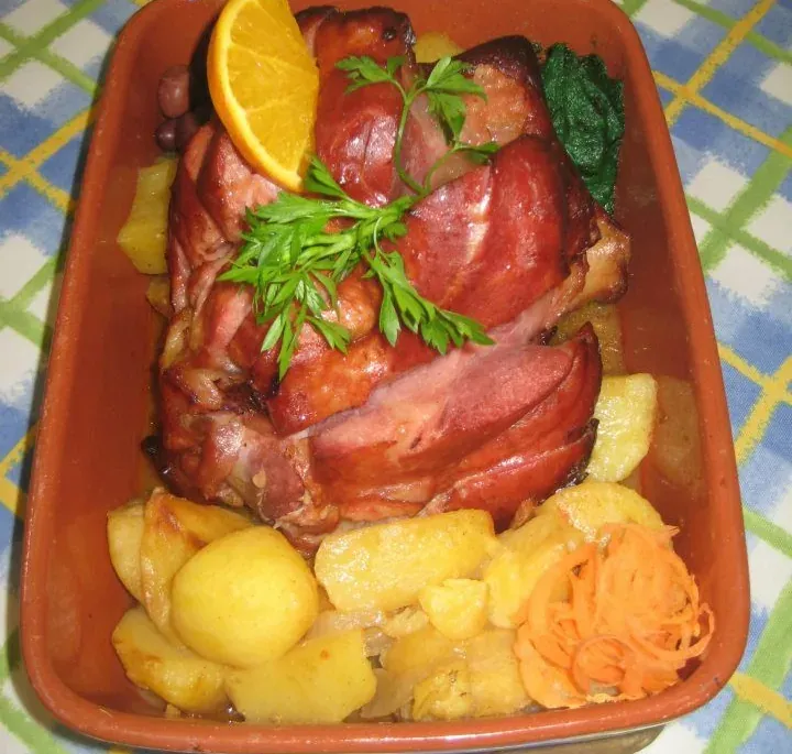Roast pork leg with potatoes and grated carrot in a clay dish, garnished with parsley and an orange slice