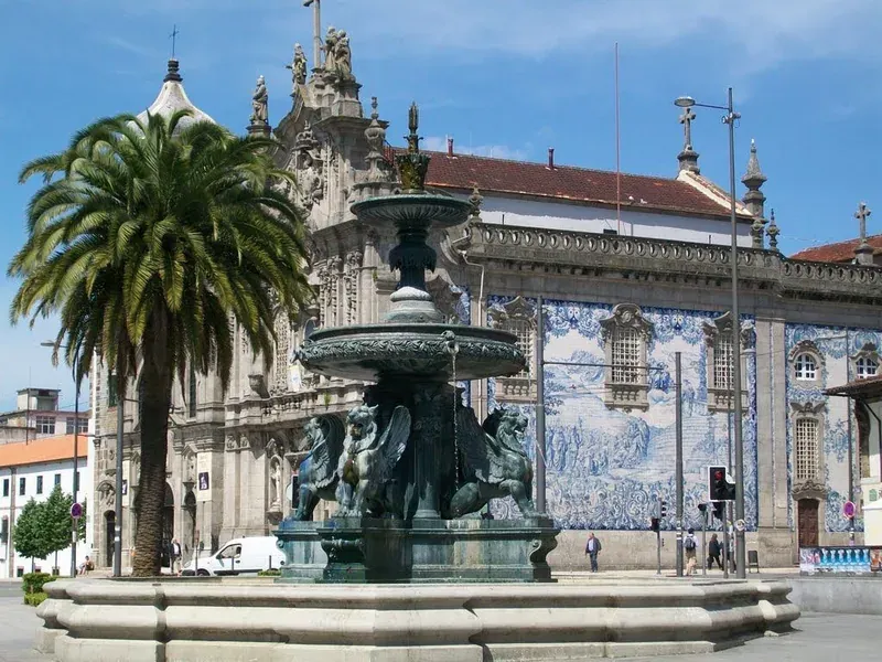 A fountain with lion statues in a square, in front of a church with a tiled side wall