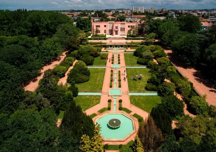 Aerial view of a pink villa at the end of a long formal garden with a fountain