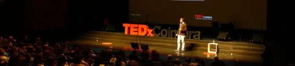 A speaker alone on the TEDxCoimbra stage, the red TEDx Coimbra letters behind him, facing a packed auditorium